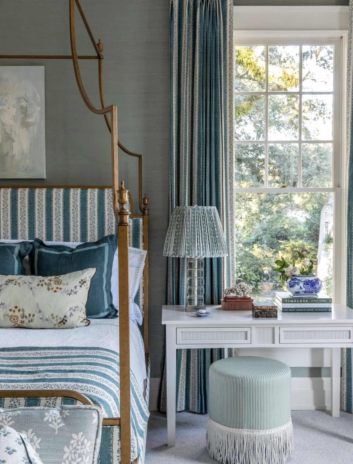 A cozy bedroom corner featuring a wooden four-poster bed with blue and white bedding, a white bedside table with a striped table lamp, a window with blue patterned curtains, and a small green upholstered bench with fringe detail.