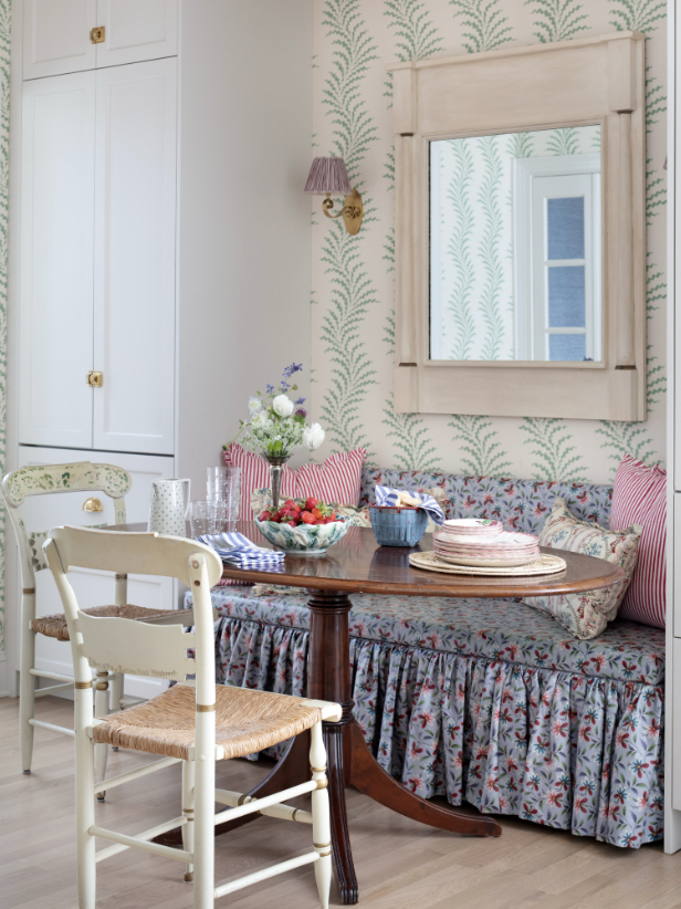 A cozy dining area with a wooden table, floral-patterned cushioned bench, and two cream-colored chairs. The table has a bowl of strawberries, a pitcher, glasses, and a flower arrangement. The wall behind has green fern wallpaper, a large mirror, and 