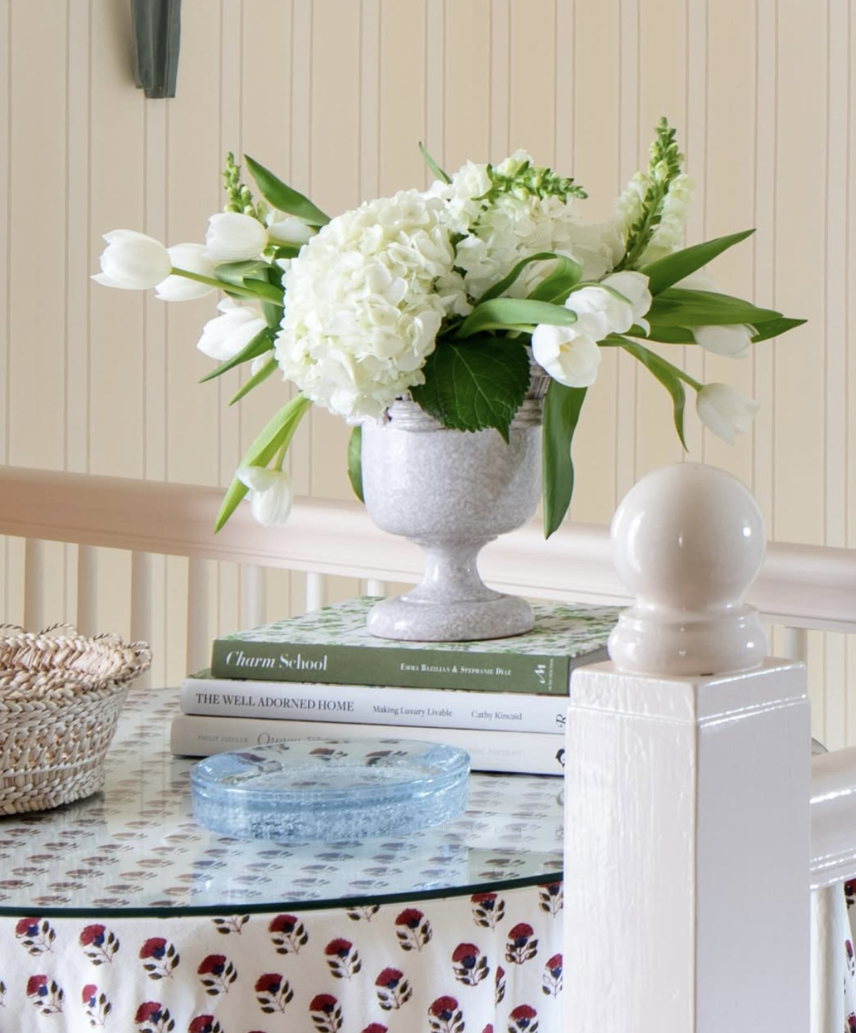 A floral arrangement with white flowers, including tulips and hydrangeas, in a gray vase on a stack of books on a table with a floral tablecloth.