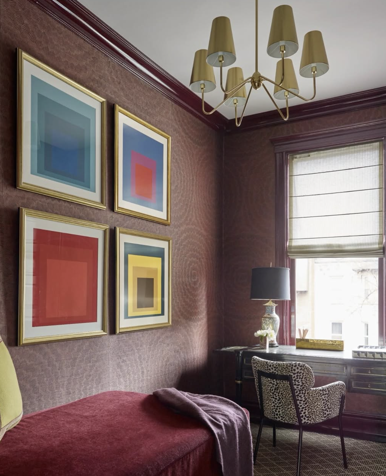A corner of a room with four framed abstract artwork pieces on the wall, a window with horizontal blinds, a velvet armchair, a table with a lamp and decorative items, and a gold chandelier.