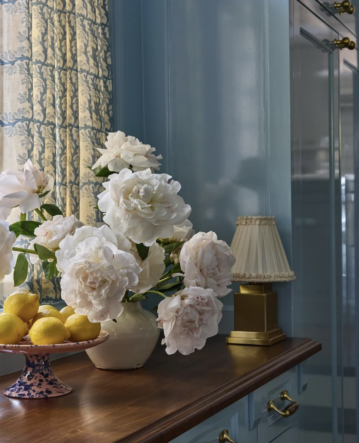 A wooden sideboard with a bouquet of white peonies in a white vase, a bowl of lemons on a pink and blue patterned stand, and a beige lampshade with a gold base, against a blue wall and window with patterned curtains.