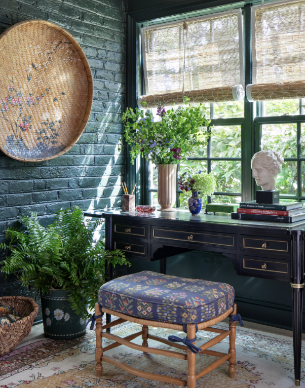 A cozy interior space with a dark green brick wall, a window with bamboo shades letting in natural light, a black desk with gold accents topped with books, vases of purple and green flowers, and a white bust sculpture. There are green plants in pots,