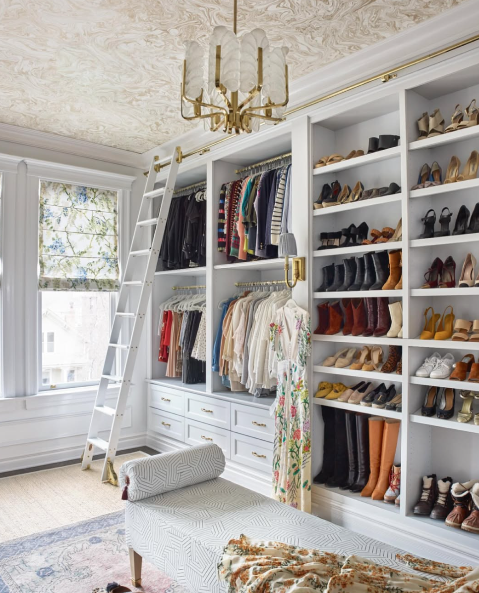 A walk-in closet with white built-in shelves and drawers, filled with hanging clothes, shoes, and accessories. A white ladder leans against the shelves, and sunlight streams through a window with a floral Roman shade. A patterned upholstered bench si