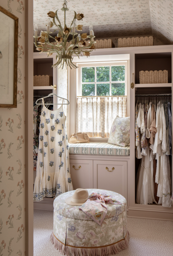 A cozy vintage walk-in closet with a window seat, floral curtains, and an assortment of hanging and folded clothes created by Jenny Holladay Design in Richmond, VA.