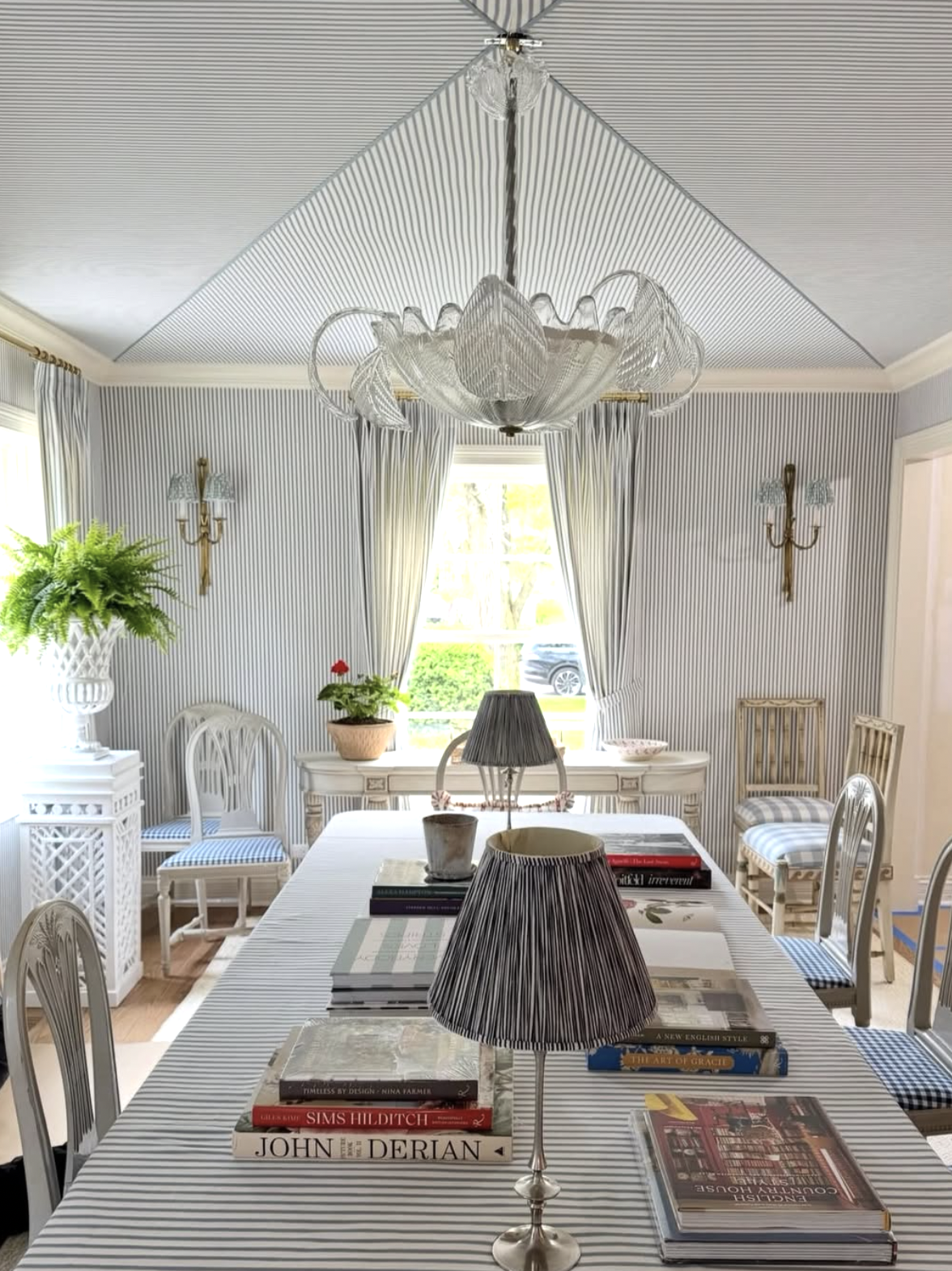 A dining room with a striped tablecloth, books, and lamps on the table. The room has striped wallpaper, a chandelier, and a window with curtains letting in natural light. There are chairs with striped cushions, potted plants, and decorative wall scon
