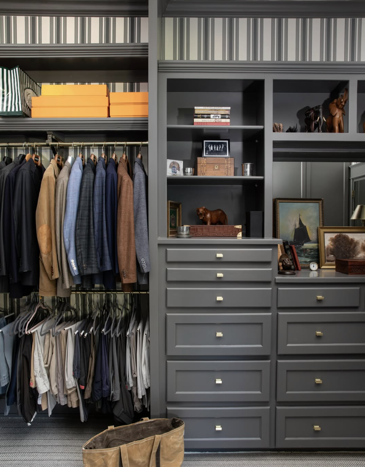 A closet with an organized wardrobe of men's suits, jackets, and shirts, with decorative items like framed paintings, books, sculptures, and a brown shopping bag on the floor.