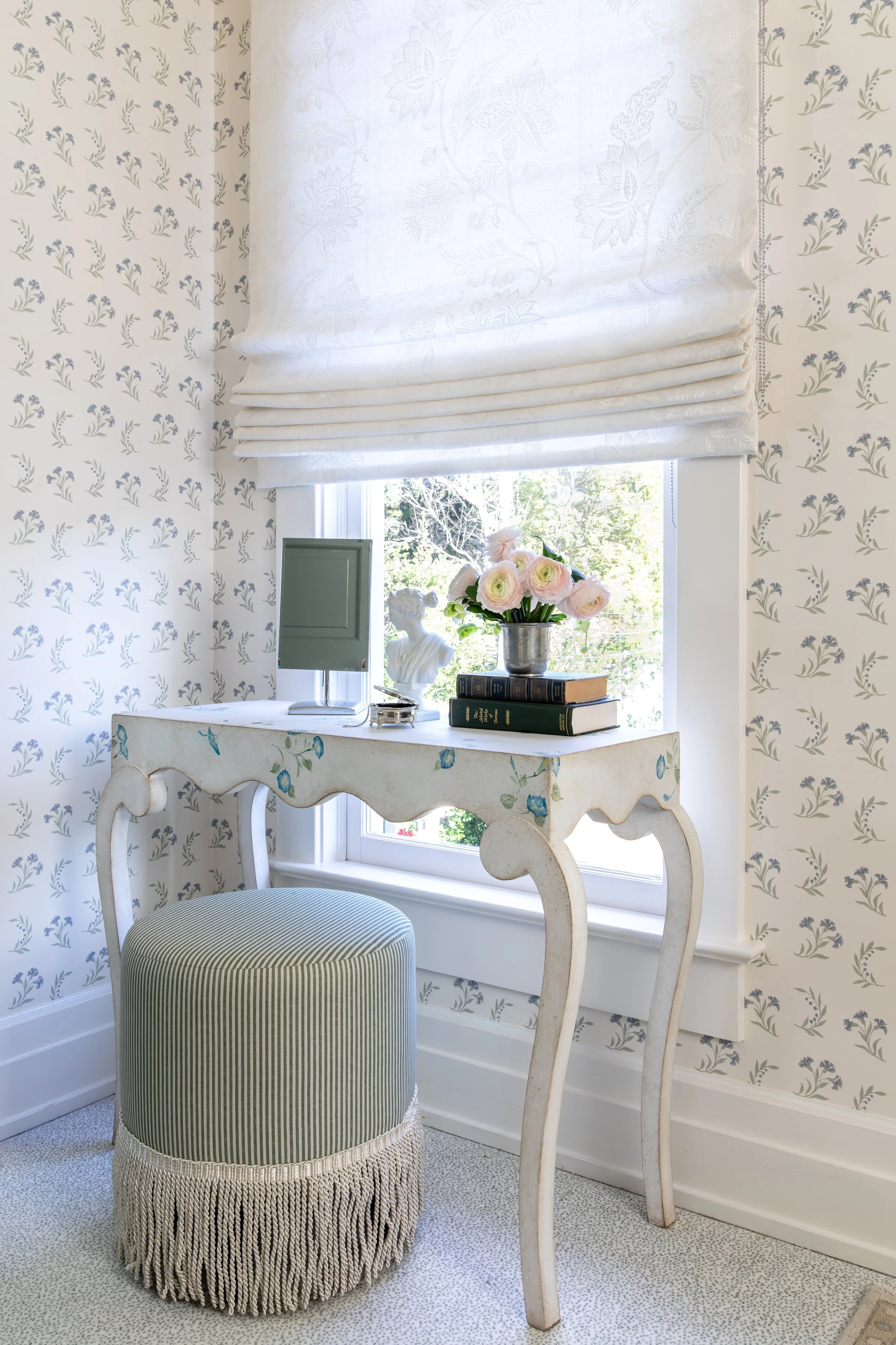 A cozy corner with a window, white floral patterned wallpaper, and a white wooden writing desk with a pale green striped upholstered ottoman. The window has a valance with a subtle pattern, and a vase of pink and white roses sits on the desk.
