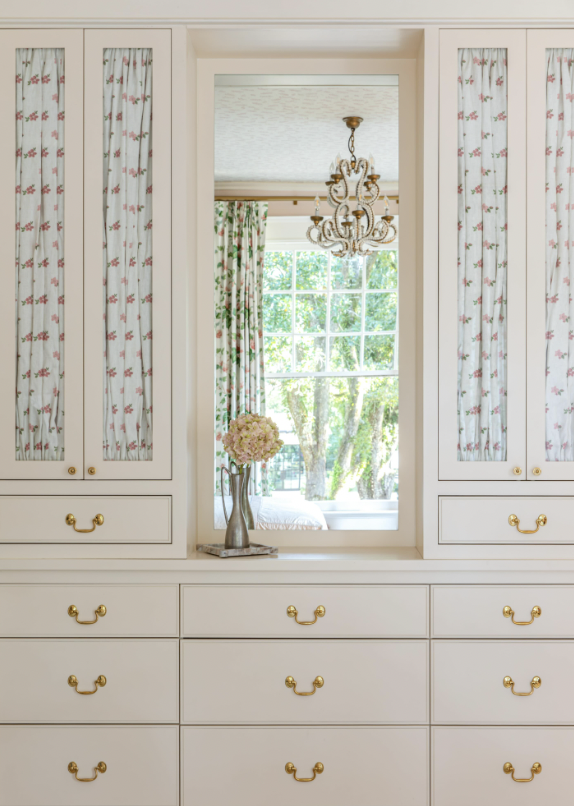A white dresser with multiple drawers and gold handles, topped with a decorative vase of pink flowers, in front of a mirror reflecting a window with trees outside and floral curtains, a chandelier hanging from the ceiling.