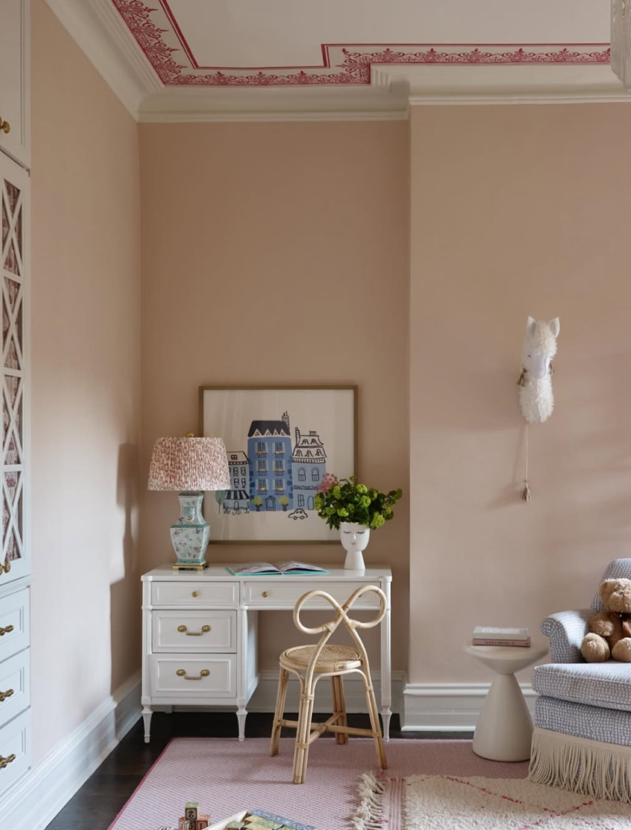 A corner of a room with a vintage white desk, a wicker chair, a table lamp with a patterned shade, a vase with green flowers, a framed artwork of buildings, a white plush llama head on the wall, a pink rug, a beige armchair with a teddy bear, and a s