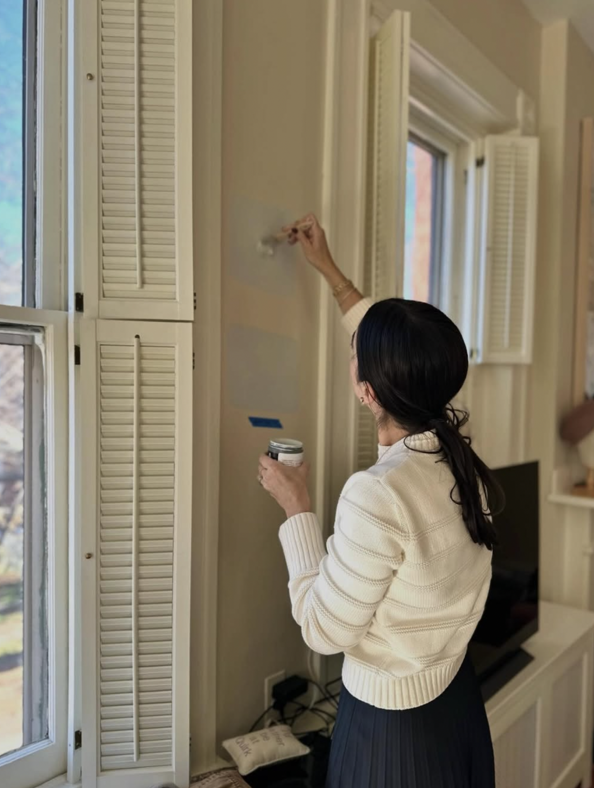 A woman painting a wall white near open windows in a living room.