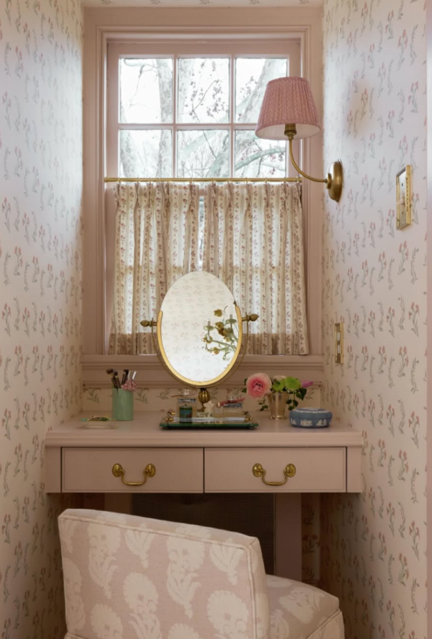 A vintage-style vanity table with a round mirror, decorated with flowers and personal items, positioned in a small, cozy room with floral wallpaper, a window with floral curtains, and a pink-shaded wall lamp.