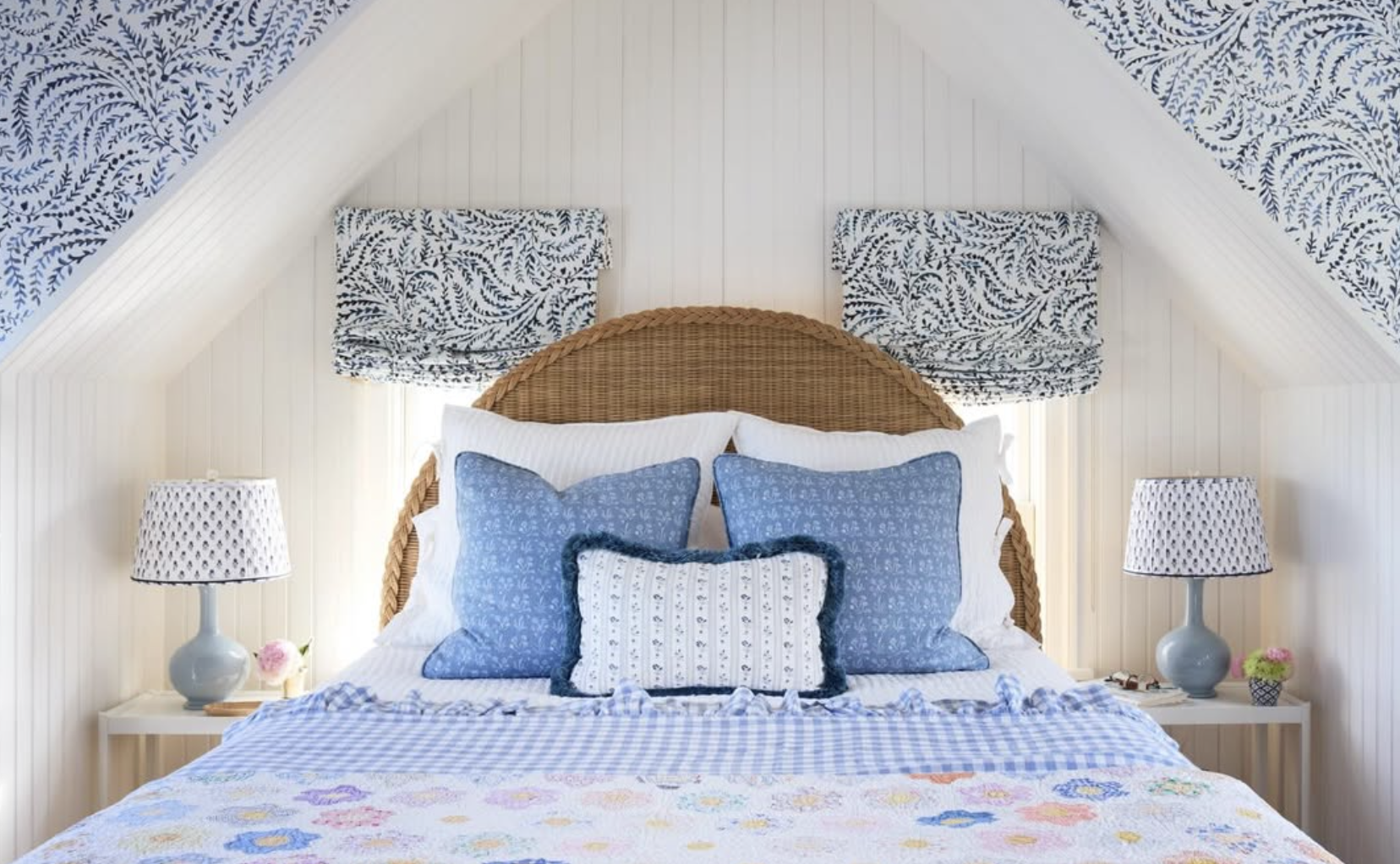 A cozy bedroom with a wicker headboard, blue and white patterned pillows, white and floral bedding, two blue lamps with patterned shades, and floral decor on bedside tables created by Richmond, VA interior designer Jenny Holladay Design.