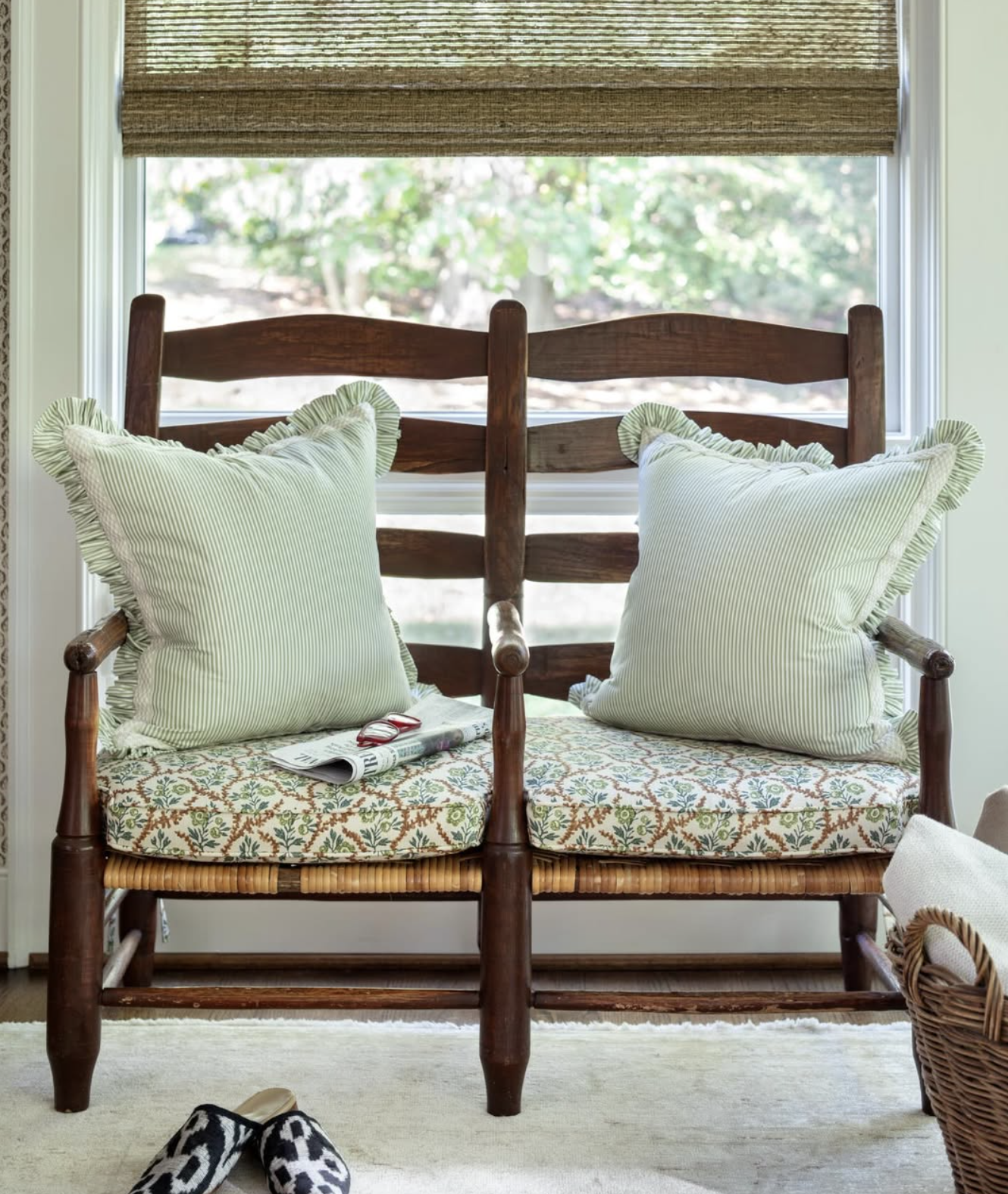A wooden bench with patterned cushions and two striped pillows positioned in front of a window with a woven shade, with slippers on the carpeted floor and a basket to the side designed by Richmond, VA interior designer Jenny Holladay Design.