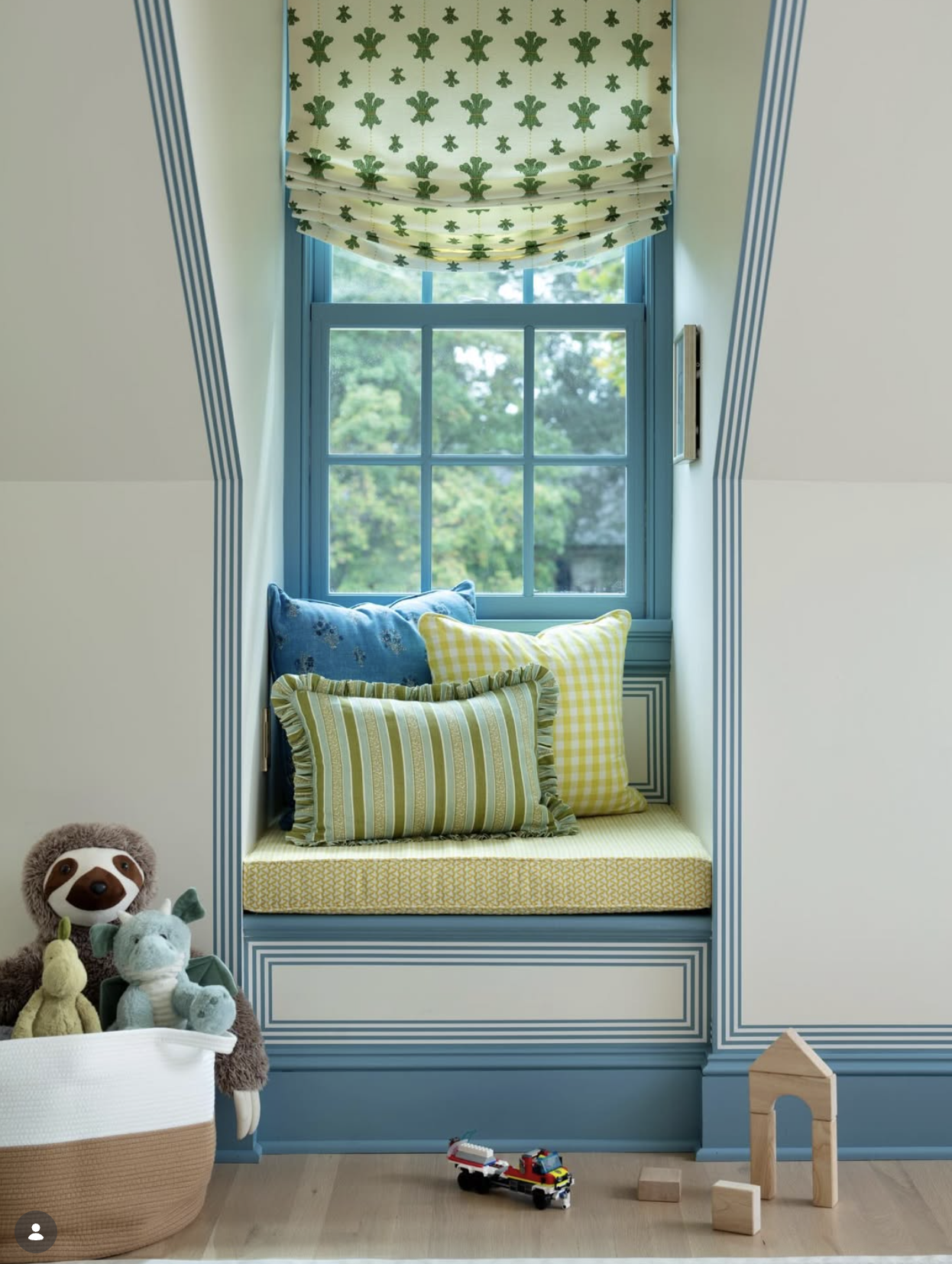 Cozy window nook with colorful pillows, a window with green foliage outside, decorative blue trim, and children's toys including plush animals, a toy firetruck, and wooden blocks.
