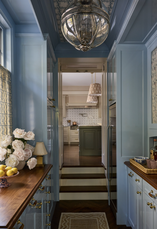 View of a bright kitchen seen through a doorway from a decorated hallway with blue cabinetry, brass hardware, floral arrangements, and a wooden side table created by Jenny Holladay Design in Richmond, VA.