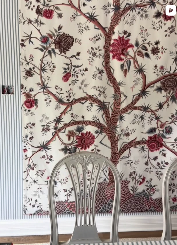 A decorative wall hanging featuring a large, intricate tree with spreading branches and large floral designs in shades of red, pink, and brown, set against a light background. Part of a dining room with silver chairs is also visible in the foreground
