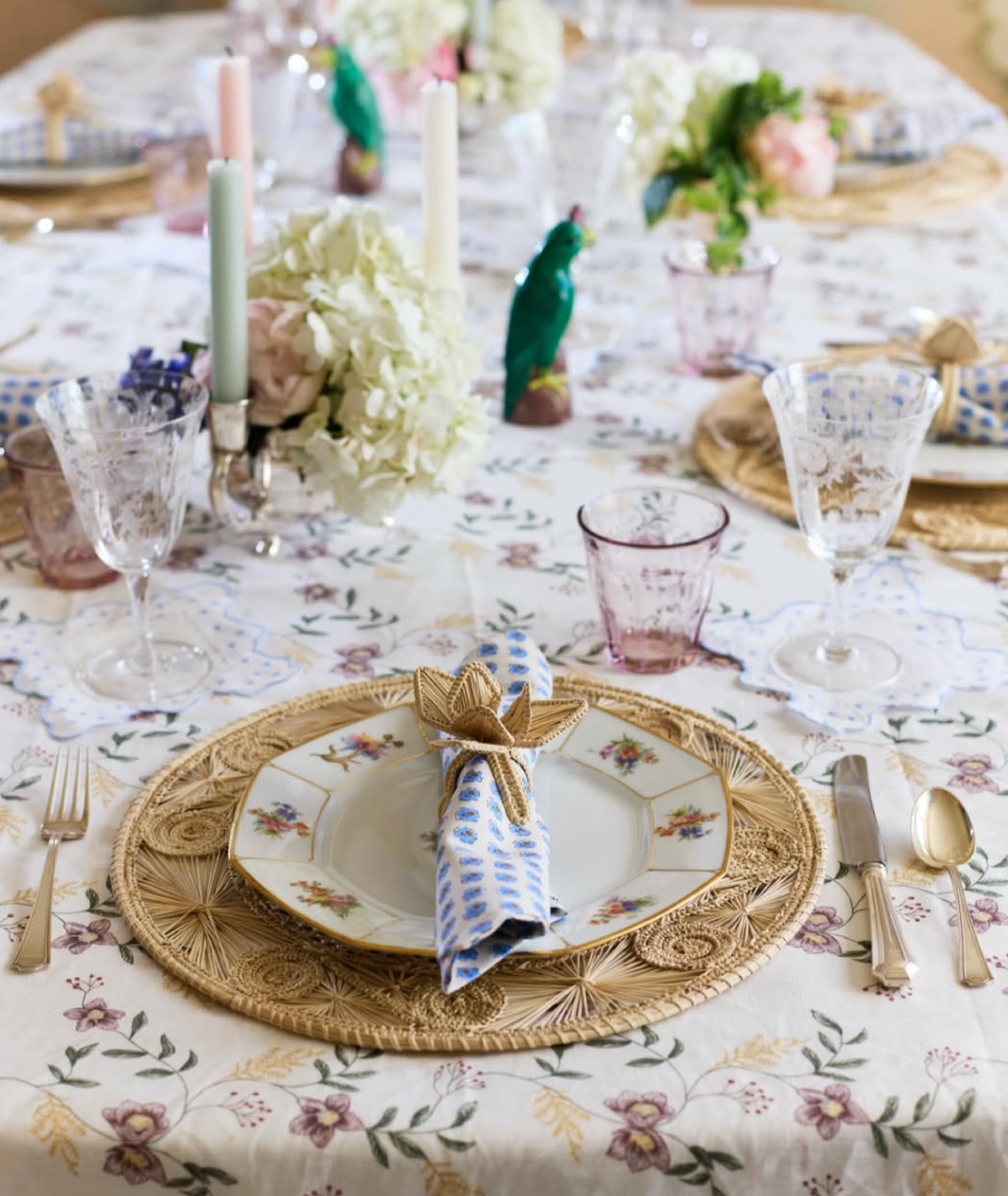 A decorated dining table with floral tablecloth, elegant china plates, gold flatware, pink and clear glasses, colorful candles, floral centerpieces, and small bird figurines.