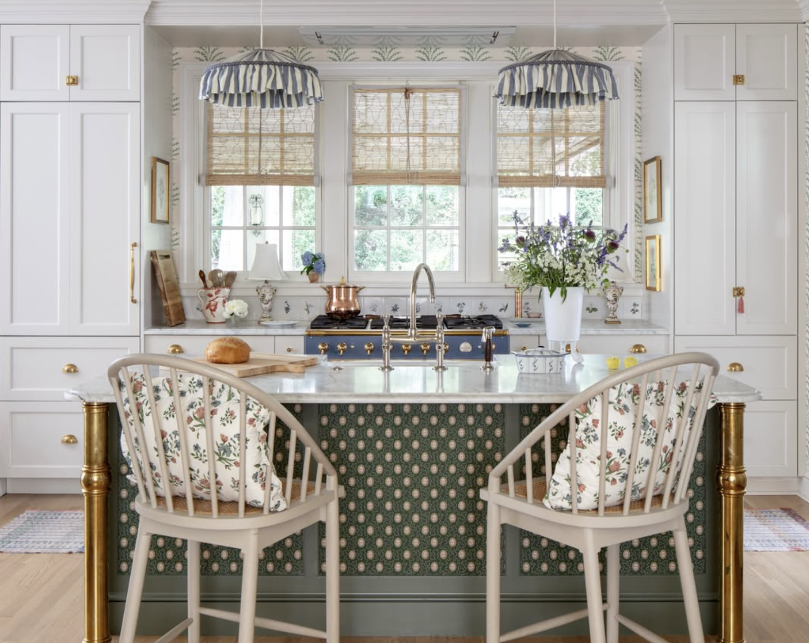 A bright kitchen with white cabinets, a marble island with gold accents, two striped pendant lights, a window with bamboo blinds, a vase with flowers, and a loaf of bread on a cutting board.