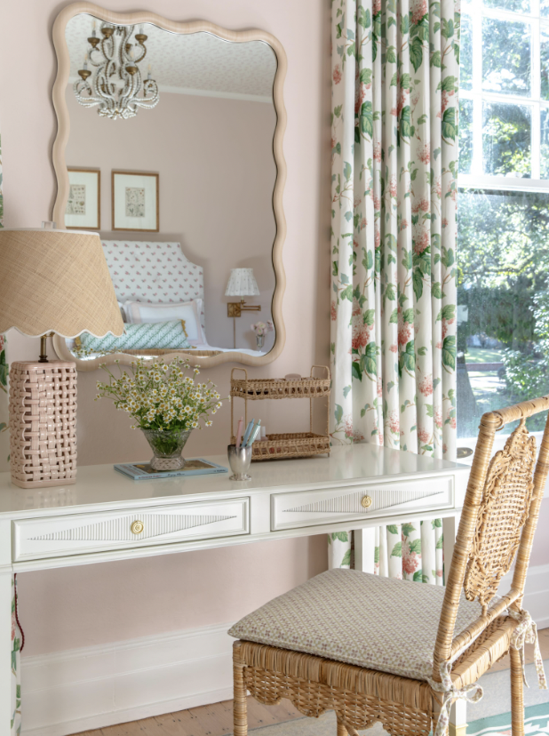 A vintage-style bedroom vanity area with a white desk, a wicker chair, a table lamp, a vase of flowers, and a decorative mirror. The scene is bright with natural light coming through a window with floral curtains. The bedroom is visible in the reflec
