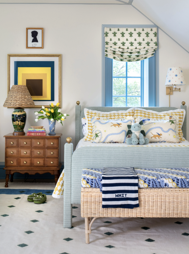A cozy bedroom with a white bed that has a yellow and white patterned duvet, dinosaur-themed pillows, and a stuffed dinosaur toy. The bed has a wicker footboard with a navy and white striped blanket labeled "WHIT." Next to the bed, there's a wooden n