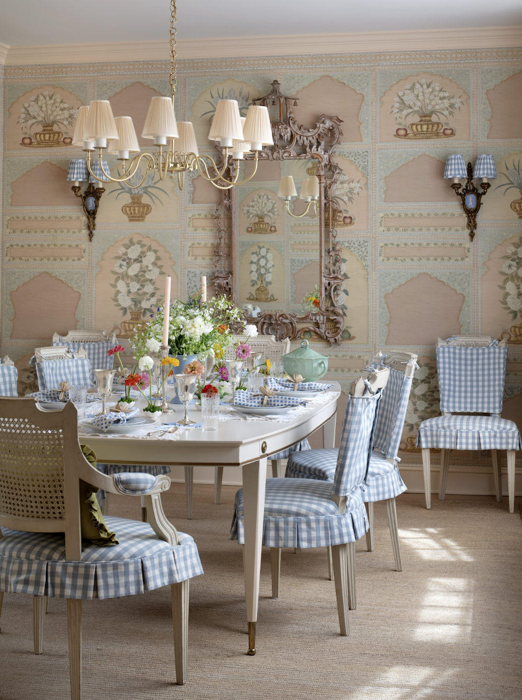 A vintage-style dining room with a white oval table set with floral arrangements, candles, and china. The room features blue and white checkered upholstered chairs, a large mirror with an ornate frame, a cream-colored chandelier, and floral wallpaper