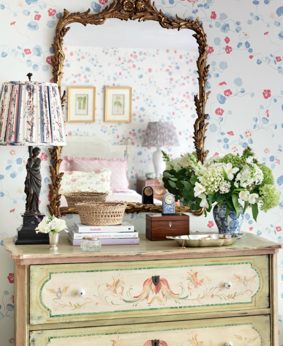 A vintage dresser with floral and bird motifs, topped with a large blue and white porcelain vase filled with white flowers, a small glass vase with a white flower, a decorative clock, a jewelry box, and a brass tray. Behind the dresser is a mirror wi