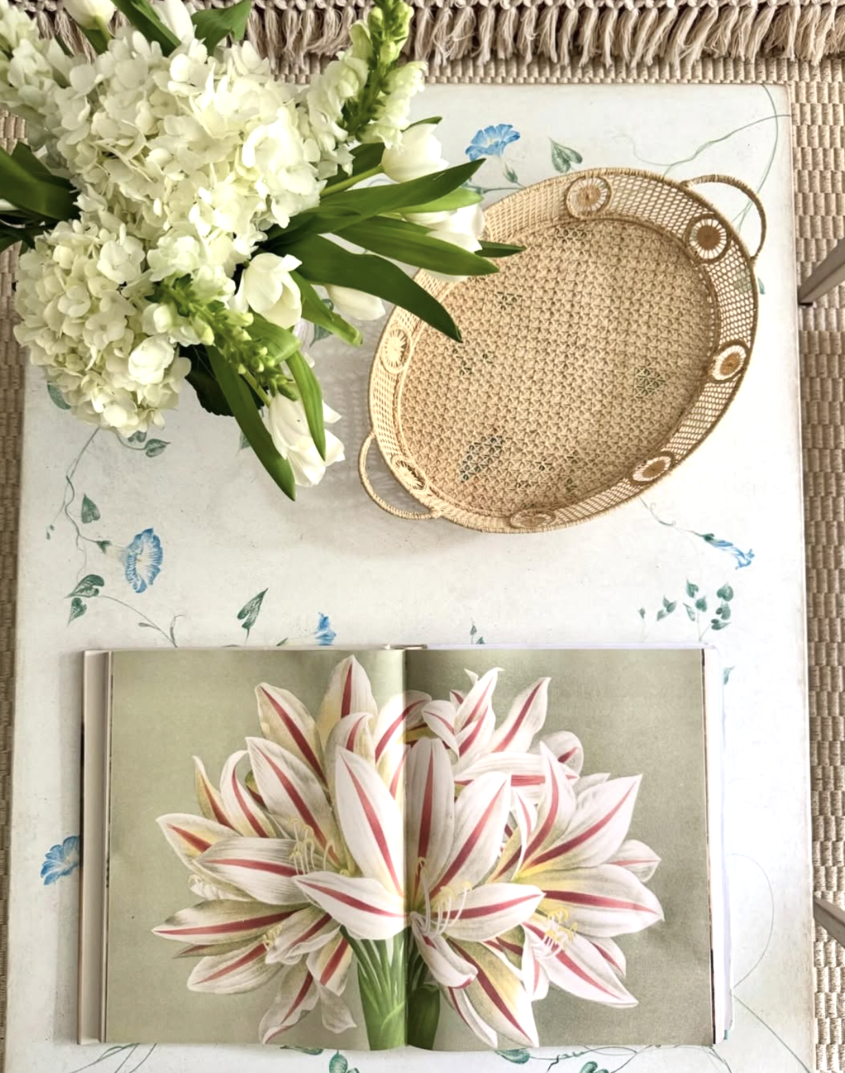 A top-down view of a table with a white floral tablecloth, a vase of white flowers, an open book with a large white and pink flower illustration, and a round woven basket designed by Richmond, VA interior designer Jenny Holladay Design.