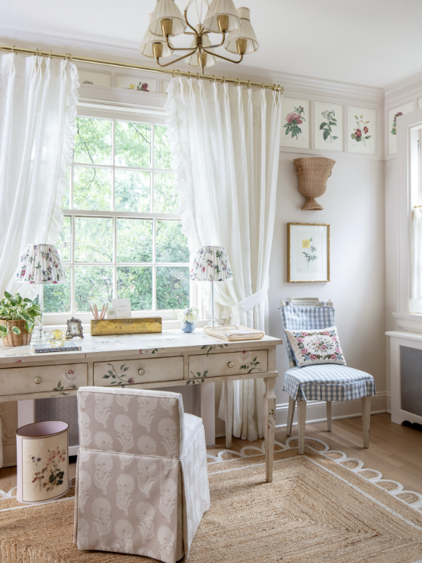 Bright, cozy room with a large window, white curtains, and floral decor. Includes a desk with plants and picture frames, a floral-patterned chair, a blue checkered chair with a floral pillow, and a woven wall basket. Warm lighting and natural light c