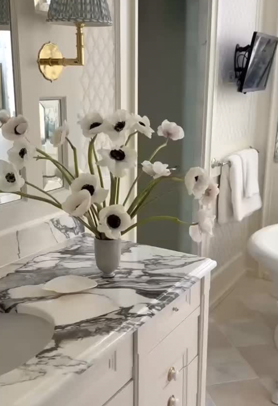 A bathroom with a white marble countertop holding a large white vase filled with white anemone flowers, a small white dish, and a decorative wall lamp. A mirror, a wall-mounted television, and folded white towels are also visible.