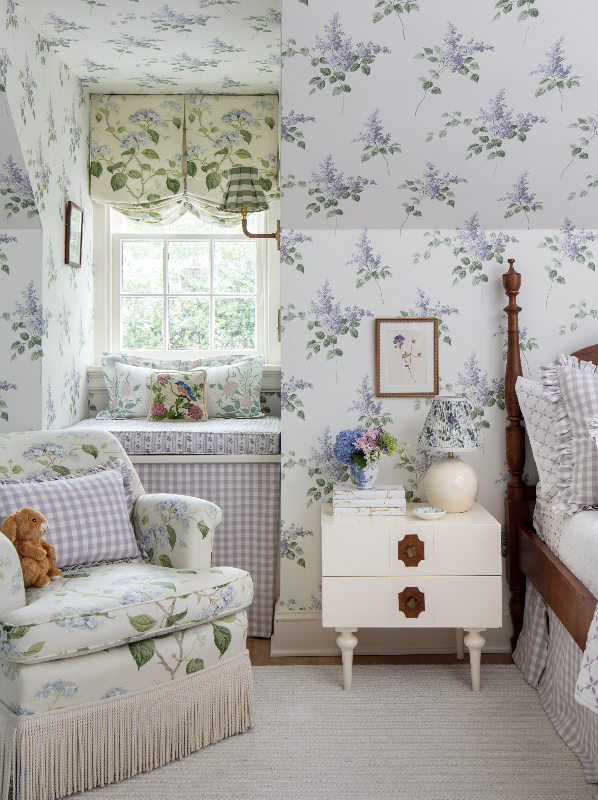 A cozy bedroom with floral wallpaper, a window seat with pillows, a floral armchair with a stuffed animal, a white nightstand with a lamp, flowers, and framed pictures.