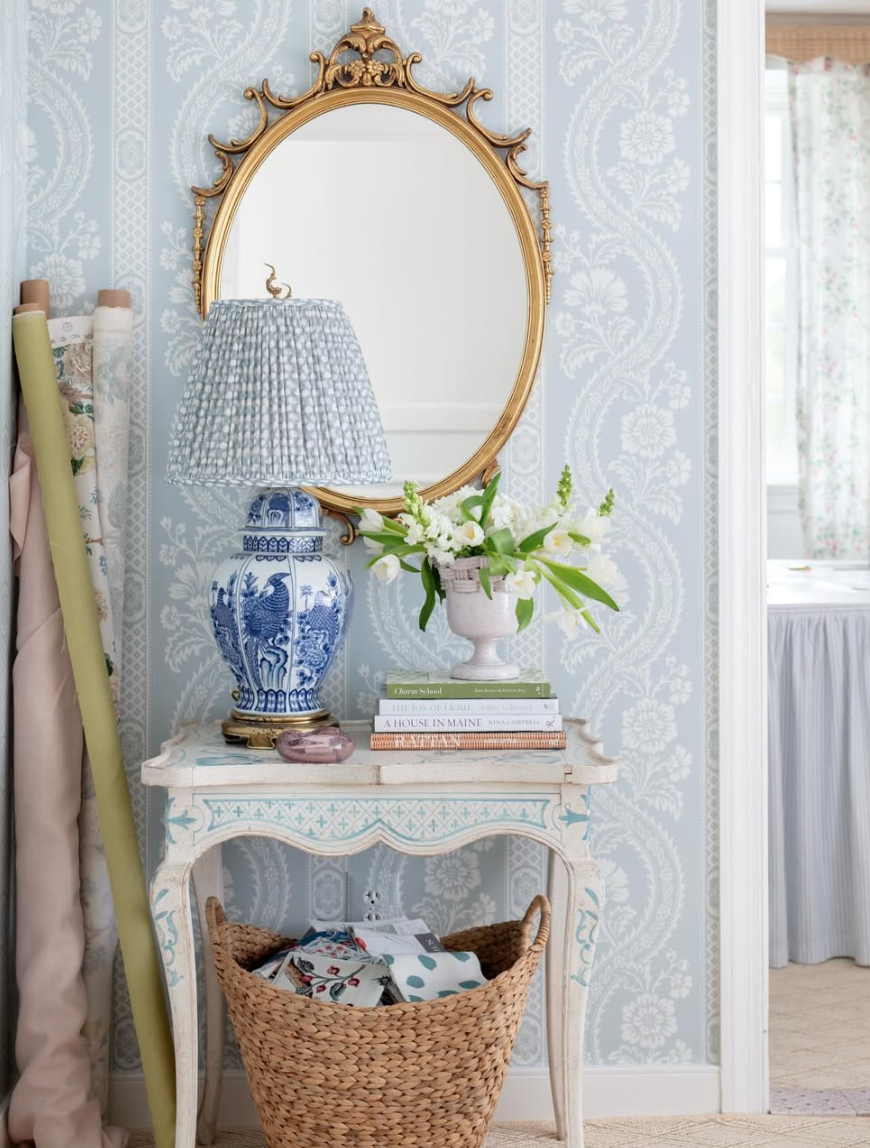 A vintage styled side table with books, a blue and white floral table lamp, a vase with white flowers, and a gold framed mirror on a patterned light blue wallpapered wall.