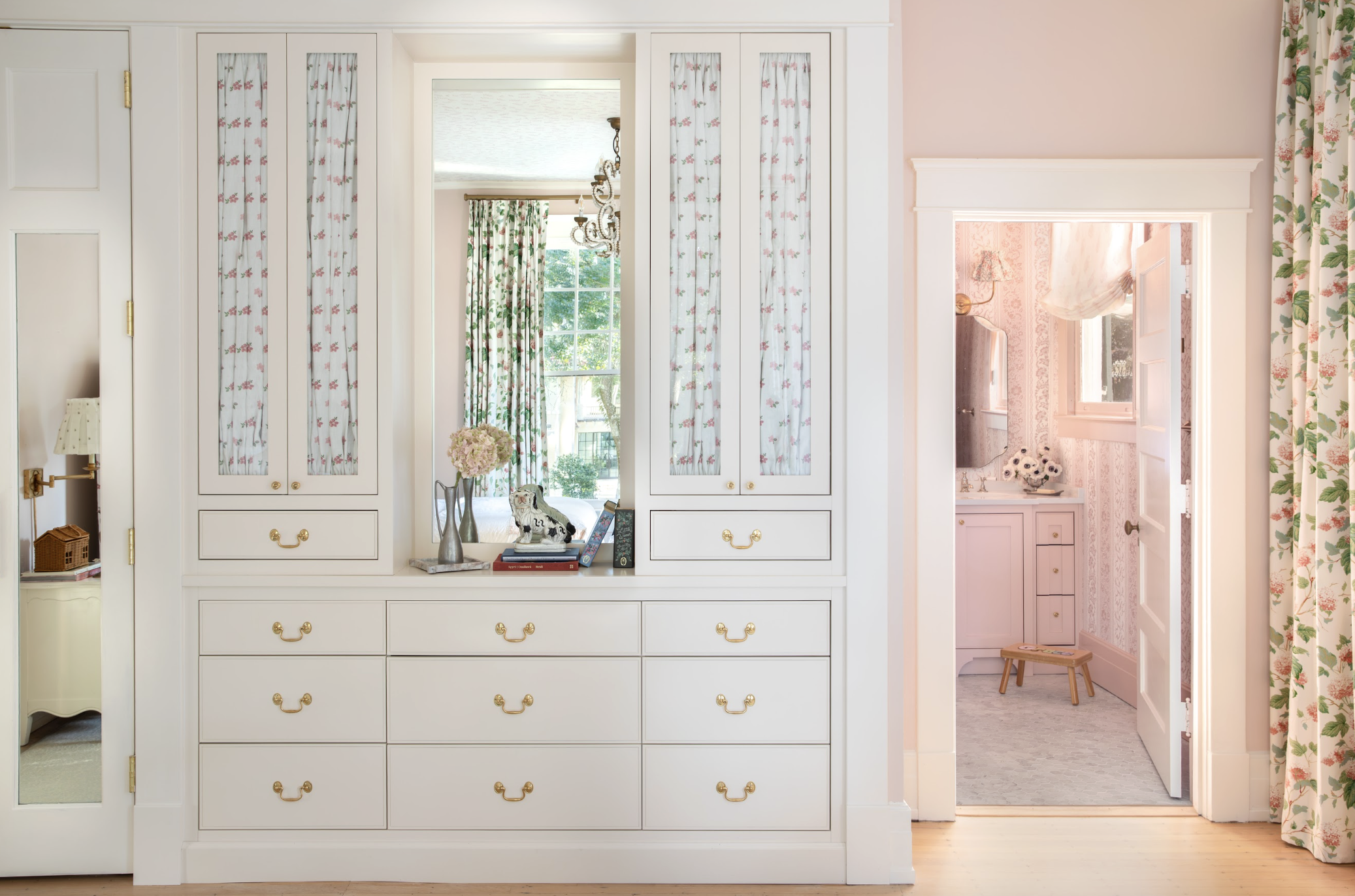 A built-in white cabinet with drawers and glass-paneled doors, decorated with floral curtains, a small potted plant, and a ceramic dog figurine on the shelf created by interior designer Jenny Holladay Design who's based in Richmond, VA.