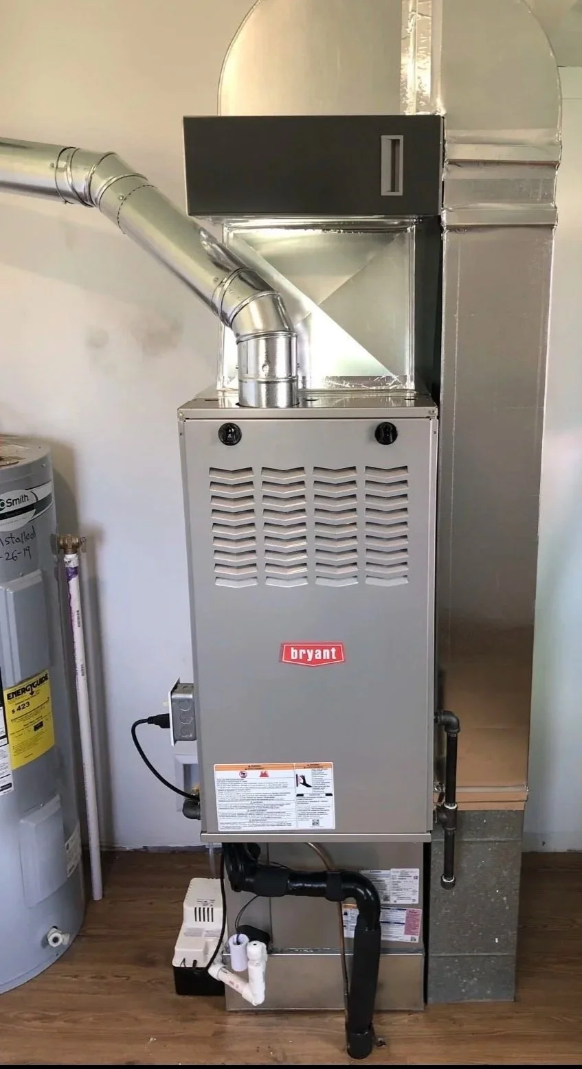 A Bryant furnace unit used for heating with metallic ductwork connected to it, located next to a water heater on a wooden floor.
