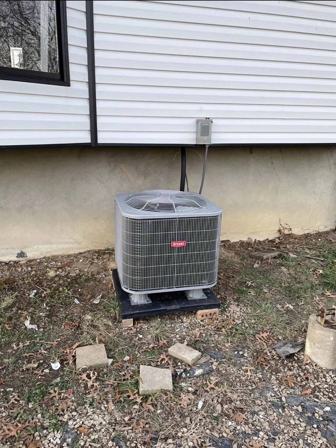 Outside of a house with white siding, an air conditioning unit installed on a black platform on ground with scattered leaves and debris. The unit has a red sticker labeled Arvant.