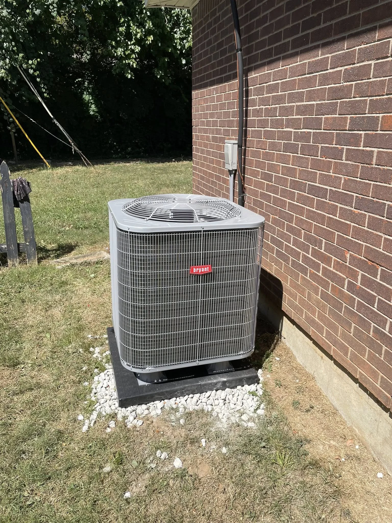 Outdoor Bryant central air conditioning unit installed beside a brick house wall, on a black concrete pad with gravel, in a grassy yard.