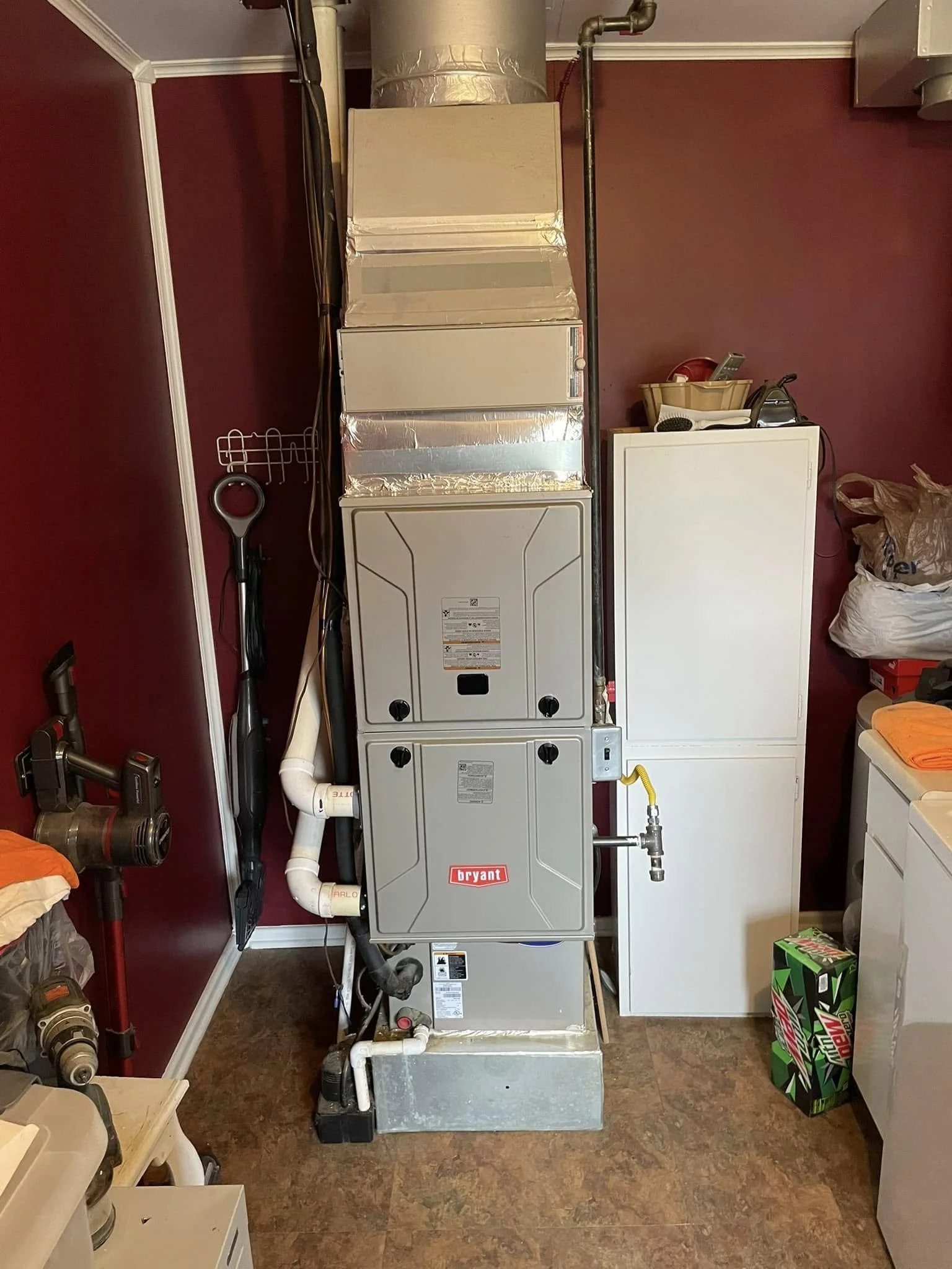 A Bryant furnace and air handler unit installed in a laundry or utility room. The unit is vertically positioned on the floor, with ductwork and PVC piping connected. To the right, there is a white storage cabinet and a stack of Mountain Dew boxes on the floor. The background features a maroon wall and various other household items.