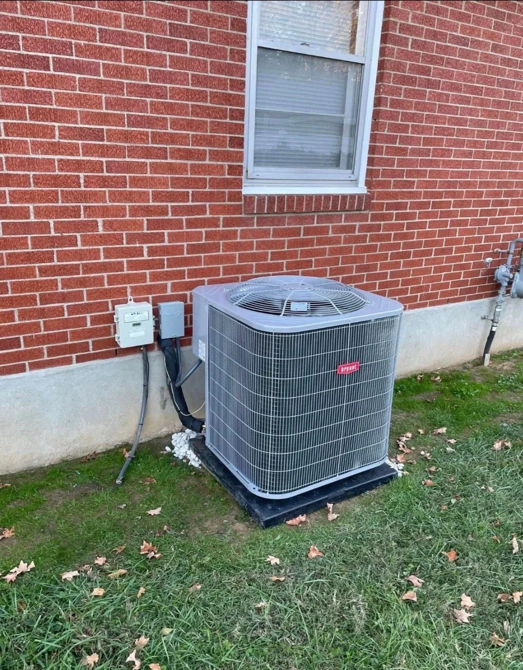 Residential building's exterior with a red brick wall, a window with closed blinds, and an outdoor HVAC unit on a concrete slab surrounded by grass with fallen leaves.