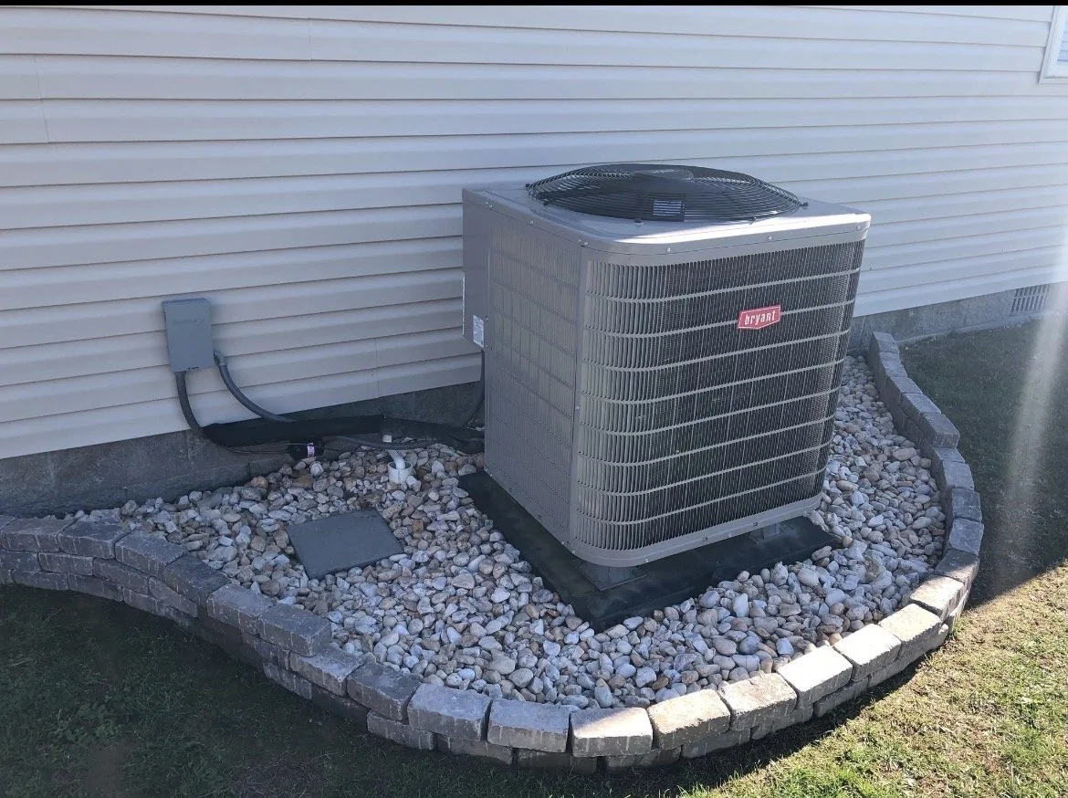 Outside air conditioning unit on a small gravel bed next to a house wall with vinyl siding.
