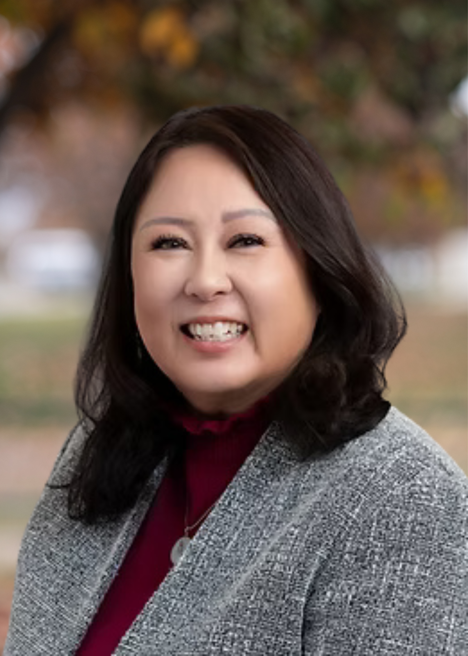 A woman with long dark hair smiling outdoors, wearing a gray blazer and a red top.