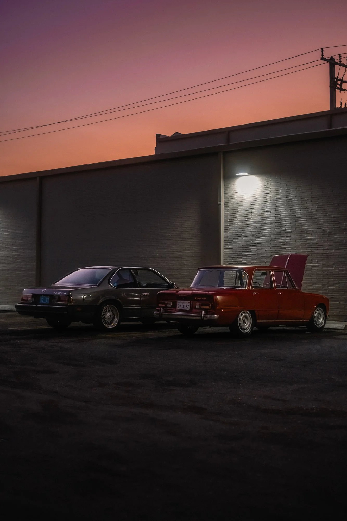 Two vintage cars parked side by side under a wall with exterior lighting, with a sunset sky in the background.