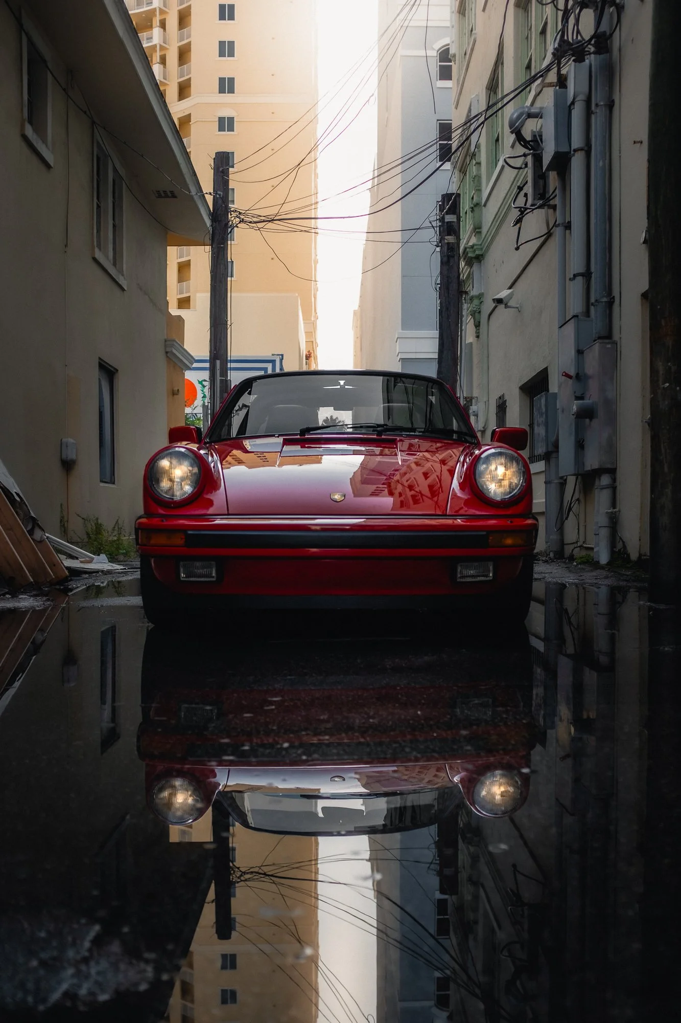 A red Porsche car parked in a narrow alleyway with puddles reflecting the vehicle, surrounded by tall buildings and utility wires overhead.