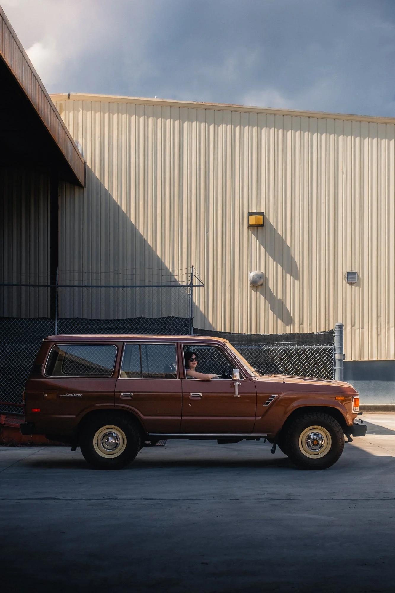 A woman with dark sunglasses sitting in the driver’s seat of a vintage brown Toyota FJ60 Land Cruiser parked at a large industrial building with a beige metal exterior wall, smiling and looking out of the window.
