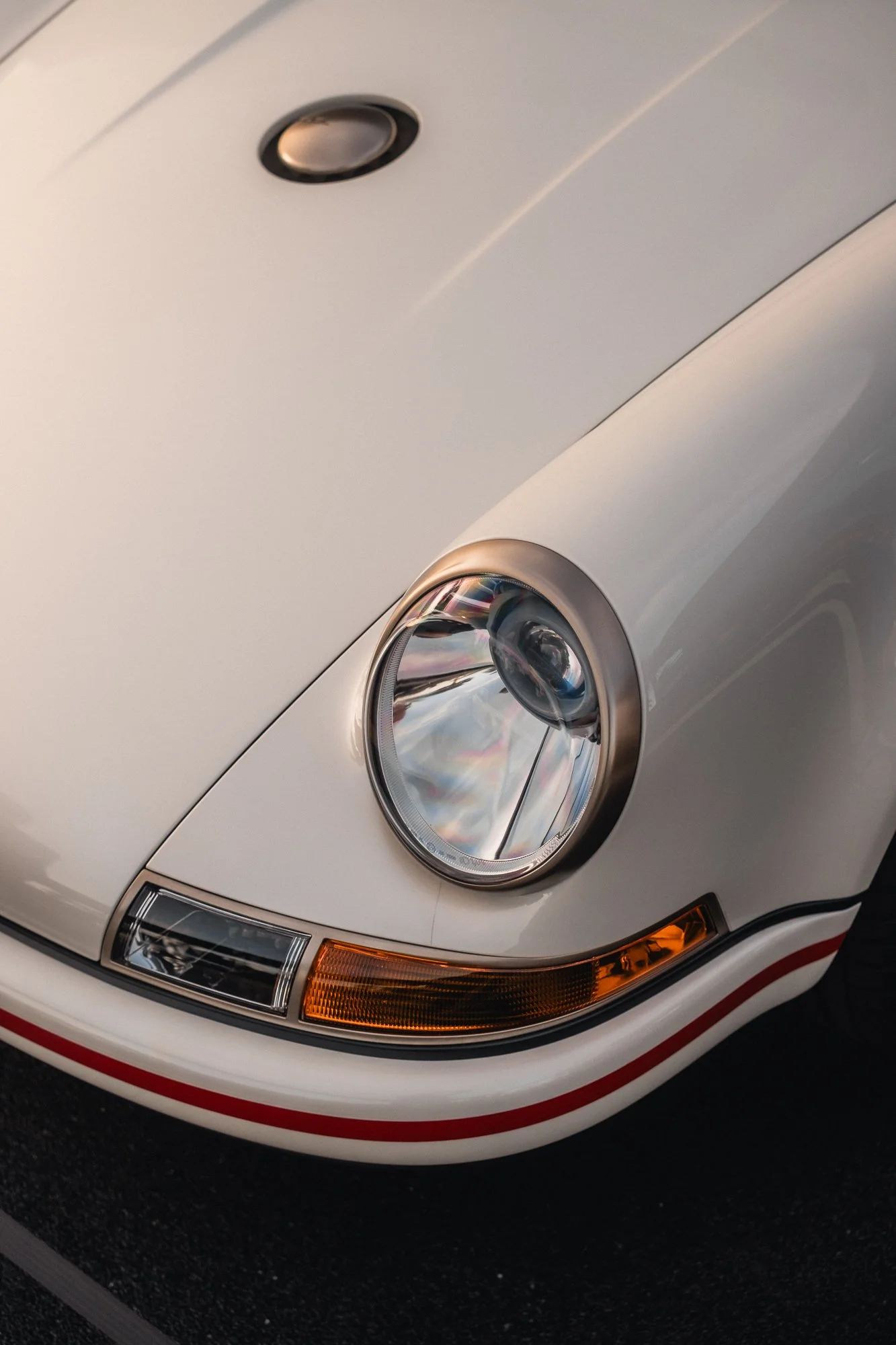 Close-up of the front of a white vintage car, showing the headlight, turn signal, and part of the hood with a red stripe detail.