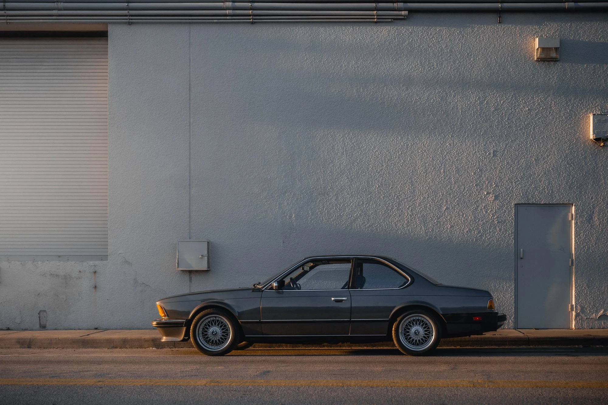 A vintage dark-colored BMW E24 633cis couple parked along a concrete sidewalk in front of a light grey industrial building with a metal door and small utility boxes on the wall.