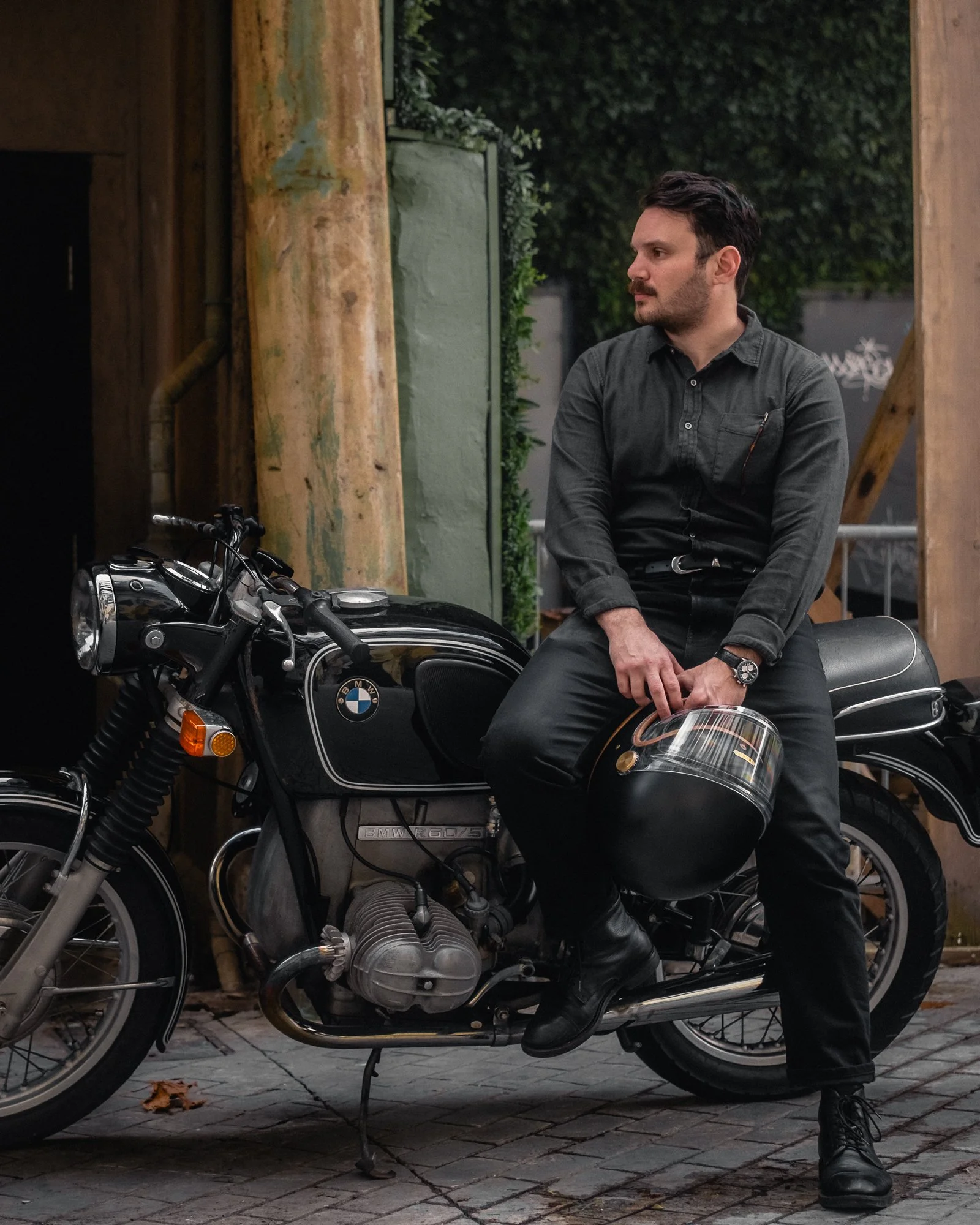 A man wearing Buck Mason holding a Hedon Workshop Heroine motorcycle helmet sitting on a vintage BMW R60 airhead motorcycle, outdoors in an urban alley of West Palm Beach, with greenery and graffiti in the background.