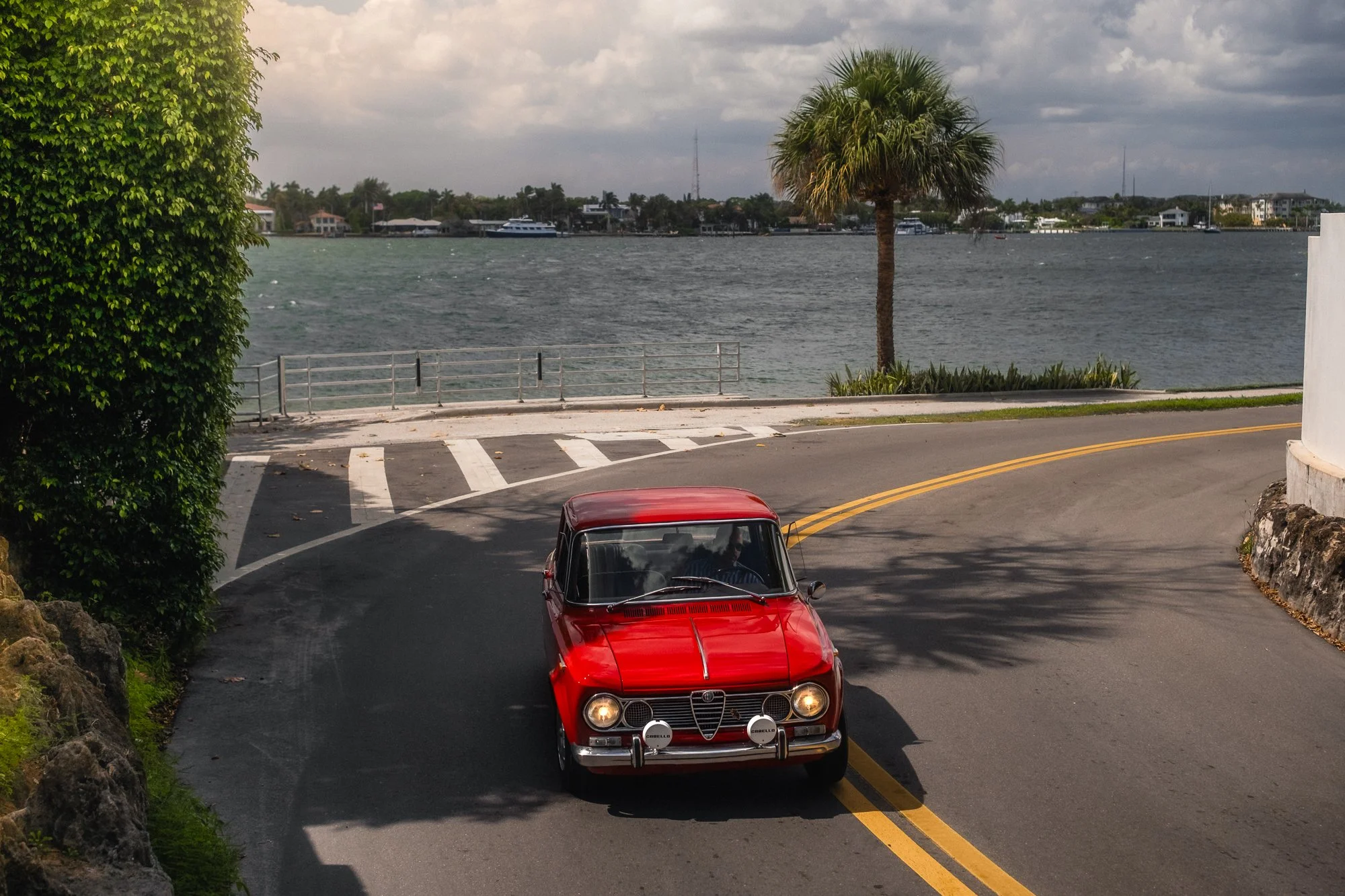 A red vintage Alfa Romeo Giulia car driving on a curved Palm Beach Island road next to an intracoastal body of water with a palm tree and boats in the background.