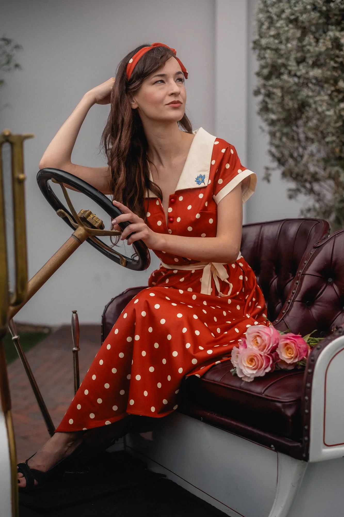 Young woman with long curly hair, wearing a red polka dot dress and matching headband, sitting on a vintage car seat inside a buggy, holding the steering wheel with one hand and resting her other hand on her head, with pink roses on the seat beside h