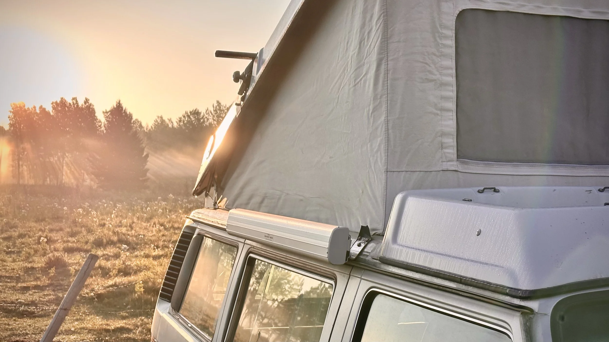 Close-up of a vintage Vanagon camper van with a pop-up roof tent set up, parked outdoors during sunset or sunrise, with trees and a field in the background.