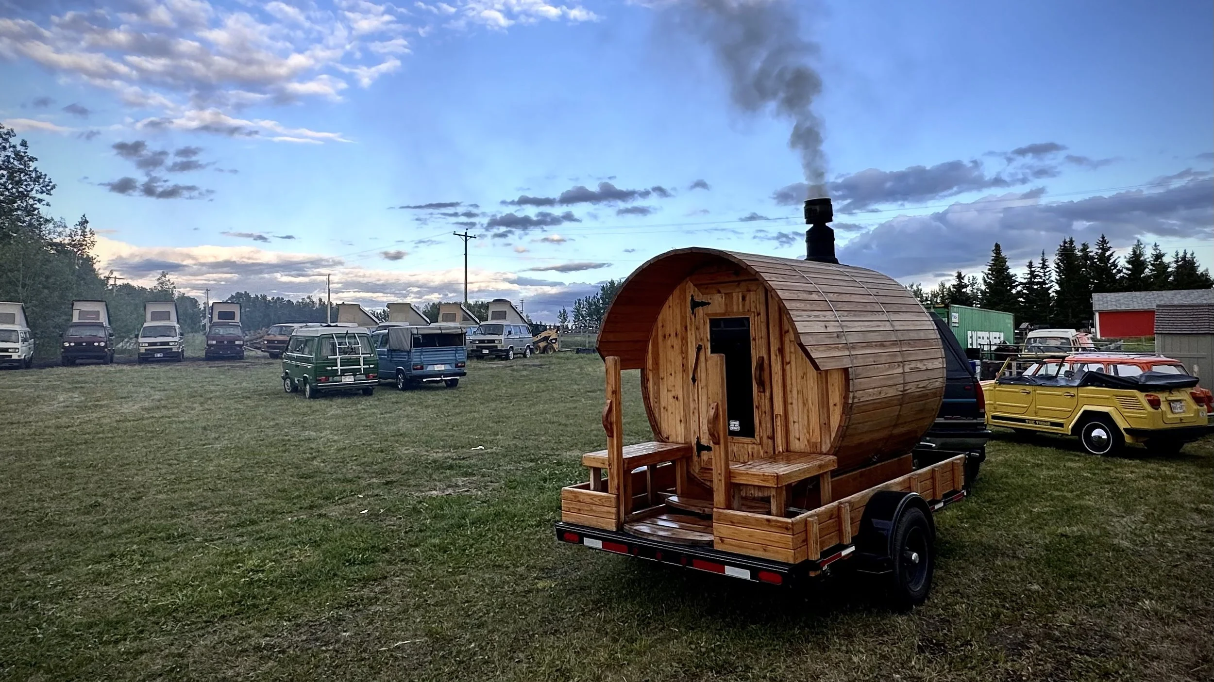 A small wooden sauna with a chimney on a trailer, parked on a grassy field with several vintage and modern cars and vans in the background, under a partly cloudy sky.