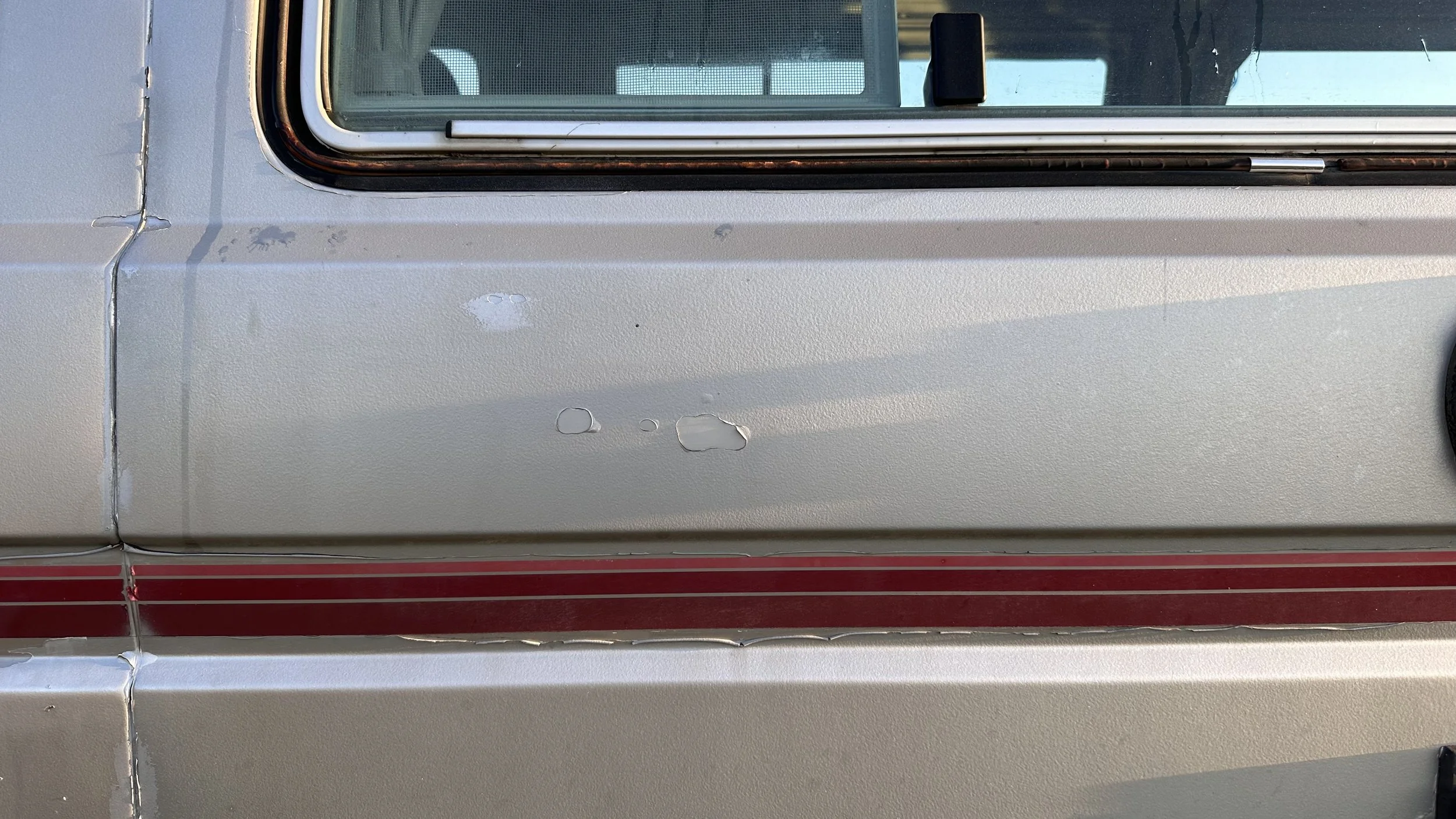 Close-up of a Vanagon exterior showing multiple dents and scratches near the window and on the door, with red and silver trim.